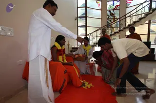 Indoor satsang house visit in a bright hall with a staircase and stained-glass windows. A woman in orange robes and a garland of yellow and red flowers sits on an orange cloth while a man in white dhoti and shirt with a yellow shawl offers a tray of fruit. A man in white shirt and dark trousers bows with folded hands at her feet, a woman in a patterned sari kneels beside him, and an older person in orange stands behind. A brass oil lamp stands nearby, and yellow marigold petals lie on the orange cloth. a person candidate is not clearly identifiable from appearance.