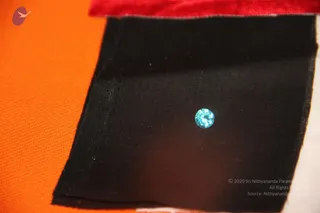 Close photograph of a single small round faceted blue gemstone. The black cloth lies on a bright orange fabric surface, with a red velvet edge visible along the top. No people are present.