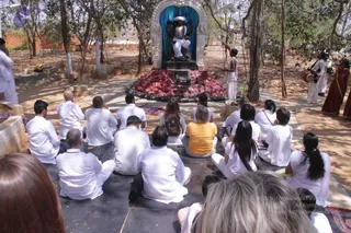 Outdoor ritual scene in a shaded garden courtyard. A dark stone deity murti stands in an arched niche with blue drapery behind it, placed on a pedestal before a mound of red fruits or offerings. A man in white stands beside the shrine. Rows of devotees in white clothing sit cross-legged on dark stone flooring facing the shrine, with a woman in a yellow top among them. On the right, drummers in white play drums and a woman in a red-orange sari stands nearby. Several people are seated and listening, and a woman in white stands at the front right. Trees and a low brick wall frame the scene.