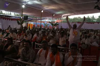 A large crowd of devotees sits in rows of metal-framed chairs under a white tent canopy at an outdoor satsang. Foreground men wear white kurtas with saffron-orange shawls, and one older man at right stands with both arms raised, wearing a white kurta and a rosette badge. Rows of seated people fill the background, with women in sarees at left. A stage backdrop with red, blue and yellow panels, green banana plants and a speaker are visible behind the audience, with palm trees beyond.