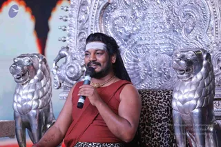 A man seated on an ornate silver throne with silver lion-head armrests and a leopard-print cushion speaks into a handheld microphone during a discourse. He has long dark hair swept back, a white vertical tilak on his forehead, a dark beard and moustache, and wears a rust-red sleeveless top with a red-and-black cloth draped over the shoulder, a gold pendant necklace, and a leopard-print lower wrap. A large silver-embossed backrest with floral and scrollwork designs stands behind him, and red and white stage lighting appears at the left edge.