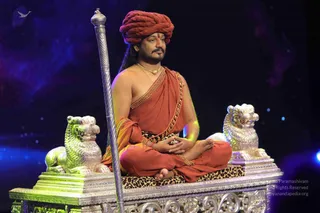 A seated man in an orange-red saffron robe and matching red-orange turban sits cross-legged on an ornate silver throne supported by two silver lion figures and a tall silver staff, with a leopard-print cushion beneath him. He wears a gold necklace, rudraksha-style beads, and a bare chest with a draped orange shawl, hands in a meditative mudra, against a dark starry blue stage backdrop. Swamiji (probable): long hair and a gold necklace with a saffron robe on a throne; appearance-based label only. Stage photograph of a single person.