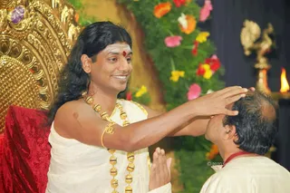 A man with long, swept-back dark hair, a red bindi and white tripundra-style markings on his forehead, wearing a white dhoti-style wrap and heavy gold rudraksha-style beaded necklaces and an armlet, places his right hand on the head of a kneeling man in a white shirt with folded hands. The seated figure sits on a red cushioned throne with ornate gold carving behind him. Behind them are arrangements of orange, pink, yellow and red flowers and a lit oil lamp on a brass stand at right. The scene suggests a blessing or darshan moment. Swamiji (probable): the long hair, gold beaded necklaces, white wrap and throne cues match the page's Swamiji reference.