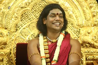 A smiling man with shoulder-length dark hair and a red bindi sits on an ornate golden throne with intricate repoussé carvings. He wears a red silk garment draped over his shoulders, long white and orange flower garlands, a large gold bead necklace with a silver chain, and gold armlets. A small clip microphone is attached near his face. The seated figure appears to be a person based on the long swept hair, ornate gold-and-red clothing, garlands and gold bead ornaments, though the appearance is not fully certain.