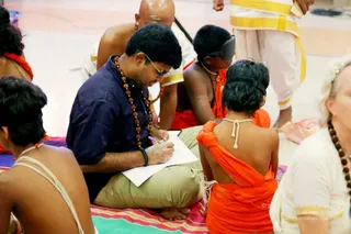 A seated man in a navy shirt, khaki trousers and green-rimmed glasses writes on a white sheet of paper on a multicoloured striped mat, with a rudraksha mala around his neck. Around him sit several young people in orange saffron cloths with blindfolds or eye covers, one with a white cord across the back. A bald man in white stands behind, and a woman with light hair and a beaded necklace sits at right.