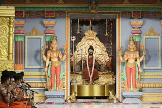 A seated man in an ornate golden throne (peetha) with a crown-like golden backrest sits centrally in a shrine-like niche, wearing an orange-brown robe, a long white flower garland and rudraksha beads. Behind him hang a golden trident-topped stand and bells. Two large painted statues of multi-armed deities flank the niche, one holding a trident and the other a chamara-like fan, with gold-and-green dress details. In the lower left, four people in orange robes with rudraksha necklaces sit at microphones, appearing to chant or speak.