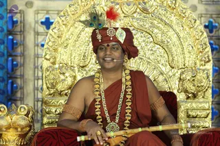 A seated man in a maroon-red wrapped turban with a peacock feather and red plume, wearing a maroon-red shawl, bare-shouldered, with layered gold and jewelled necklaces and gold armbands. He holds a bamboo flute across his lap and wears a small headset microphone. Behind him is an ornate gold throne with carved floral relief, and a gold lion-like armrest sits at the left edge. Appearance-based label: a person, seated on the golden throne and speaking as a discourse presenter, with peacock feather, plume and jewelled turban cues.