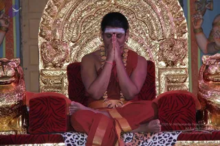 A photograph of a seated man with a shaved head and a white vertical tilak on his forehead, palms pressed together in namaskar and eyes closed. He wears a red-orange unstitched robe with a gold border, a gold-and-beaded necklace, and a pink wristband. He sits cross-legged on a red-and-black striped cushioned throne with a gold-embossed backrest and gilded lion heads on either side. A leopard-print cloth lies beneath him. Gilded figures and a decorated pillar appear at the edges. The page flags a person at this event, but the appearance cues are not distinctive enough to confirm identity, so the person is described by role.