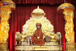 A man seated cross-legged on an ornate gilded throne with lion figures and a leopard-print cushion on a red-draped stage, speaking into a microphone on a stand. He wears a rust-orange turban, orange-red robe, long gold-beaded mala necklace, and gold bracelets, with eyes closed and a smile. Probable Swamiji by appearance: a turban, saffron-orange clothing and ornate gold necklace on a throne, consistent with a live satsang discourse. A small side table holds a card, and gilded lion-pillars flank the throne.