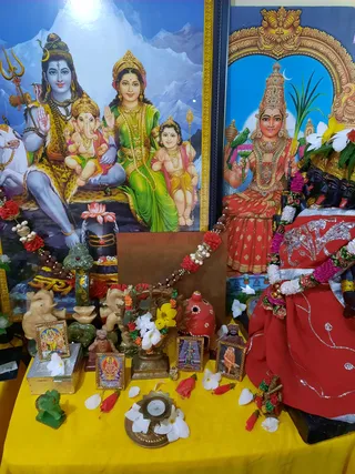 A home shrine on a yellow cloth with a large framed print of Shiva seated with Parvati, Ganesha and Murugan, a white bull at left, and a snowy mountain backdrop. Beside it stands a framed image of a goddess in red and gold holding a parrot and sugarcane. A black stone deity murti is draped in red fabric with silver embroidery and garlanded with flowers and rudraksha. Small framed pictures, a green stone Ganesha, brass lamp with a lit candle, scattered white and red flowers, and a red kalasha-like pot sit on the table.