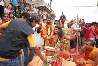 Outdoor temple scene at a stone-carved shrine, with a crowd of onlookers behind a low barrier. A central figure with long, swept-back dark hair sits on an ornate golden throne wearing an orange-saffron garment, layered beaded necklaces and gold ornaments, extending one arm forward over a brass plate and small brass puja vessels on a red-and-orange cloth. To the right, a man in orange robes holds a tall silver trident (trishul). A man in an orange-and-white robe with a flower garland stands beside the throne, and a shaven-headed man in orange with a rudraksha mala kneels at the right edge. A man in a dark cap and yellow shirt bends in the foreground, handling offerings. Visible people include the seated figure, several attendants, and numerous devotees in the background.
