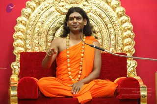 A seated man in orange saffron robes sits cross-legged on a red velvet throne with gold-embossed backrest and armrests against a red wall. He wears a long gold bead necklace, a red tilak on his forehead, and has long swept-back dark hair, and he gestures with one hand while speaking into a microphone on a stand. The ornate gold throne behind him is decorated with scrolling floral carvings. Cues on the person are long hair, saffron garments, gold beads and seated teaching posture, consistent with the page's Swamiji reference, which is an appearance-based candidate only.