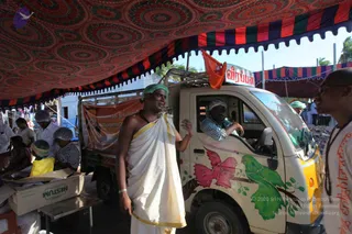 Outdoor relief camp under a large patterned canopy with red, green and navy striped valance. A man in a white dhoti with a yellow border and a rudraksha mala stands in front of a small white truck painted with pink and green birds and leaves, with an orange flag on its roof. Another man in a green head cover sits in the driver's seat. Volunteers in white head covers and green caps work at tables with cardboard boxes and yellow bags to the left. A sign on the truck reads Veerappan.