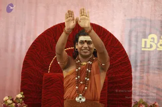 A man in orange saffron robes sits on a red velvet throne-style chair with a tiger-striped pattern, arms raised with palms facing forward in a blessing gesture. He wears a white vibhuti line and red kumkum on his forehead, long dark hair, gold earrings, multiple rudraksha malas, a beaded necklace with silver-tone beads, and a silver ornament at his chest. Bangles of rudraksha beads are on his wrists. Pink draped backdrop, a yellow Tamil-script sign on the right, and pink and white flower arrangements in the lower corners. Long hair, saffron clothing, rudraksha malas and ceremonial jewellery match the probable Swamiji description, presented in a satsang setting.
