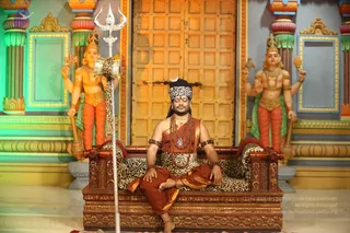 A seated man in ochre and saffron robes sits cross-legged with eyes closed on a carved wooden throne draped with a leopard-print cloth. He wears a white headband with a crescent emblem, layered rudraksha malas, a pendant, and gold armlets. A silver trident stands beside him. Behind him, two painted deity murti figures with multiple arms stand flanking a large ornate wooden door, with colourful pillars and painted walls around them. Probable Swamiji: the seated man shows long dark hair, a rudraksha mala and ornate ceremonial clothing, so the figure is. The scene is a staged darshan-style setting with a trident and a throne.