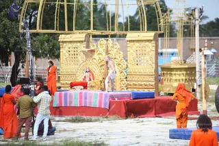 Outdoor photograph of a mallakhamba-style pole yoga set-up on a paved ground beside a gilded golden canopy and ornate golden throne-like structure. A bare-chested man with long hair stands beside a tall wooden pole, reaching toward it, in white shorts. Behind him a seated figure in red and gold sits on a golden throne with a red cushion, and a striped cloth covers a raised red platform with blue mats. Several people in saffron-orange robes stand around, including a woman in orange at left and an attendant in orange at right, while camera operators in casual clothes and a camera rig are visible at left. Palm trees and a golden chariot-like structure appear in the background.