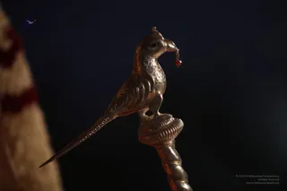 Close-up photograph of a gilded metal bird finial atop a twisted staff, shown against a dark background. The bird, with a long curved tail and a small red bead hanging from its beak, perches on a ribbed circular base. A blurred cream fabric with red stripes appears at the left edge. No people are visible.