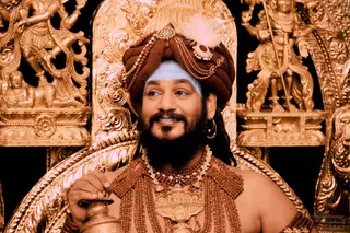 Close-up photograph of a bearded man seated before an ornate gilded throne with carved figures of deities in the background. He wears a brown turban with a white feather, gold chain ornaments, a white forehead marking, and layered rudraksha malas and beaded necklaces with a gold pendant over a bare chest. He holds a small brass kalasha vessel in his right hand. a person: long dark hair, saffron-brown turban with gold ornaments, and rudraksha malas match the page's presentation cues, though the appearance is only partly distinctive.