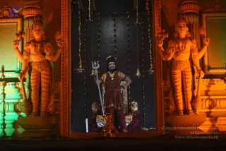 A photograph of a figure standing in a lit alcove between two large sculpted guardian statues with raised arms, holding a trident. The central person wears a maroon garment, a red-brown rudraksha-style bead mala, a gold necklace, and a crown, with dark face paint, and stands before a dark draped backdrop strung with small lights and hanging bells, with a golden throne behind. The statues on either side are orange-toned with carved jewellery and raised hands holding implements, and a brass lamp stands at left. The image carries a copyright line in the lower right.