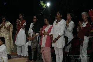 Night outdoor scene of women standing in a row with palms pressed together in anjali mudra, appearing to take part in a devotional gathering. From left: a woman in a pale gold sari with a rudraksha-style necklace, a woman in white kurta and pants with a red-orange shawl, a woman in white with black-framed glasses and a patterned grey-black shawl holding a small paper, a woman in pink top and white dupatta holding a booklet, a woman in white tunic with a gold necklace, a woman in white with a lanyard and a small paper, and an older woman with grey hair in white with a red shawl. A lamp post glows in the dark background, with dark chairs at right and a seated person in white at far left edge.