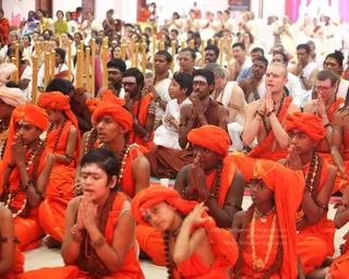 A crowded indoor satsang hall where many people sit on the floor in prayer. In the foreground, rows of young men and boys wear saffron-orange robes and orange turbans, with rudraksha malas around their necks, palms pressed together in anjali. Behind them, devotees in white and cream clothes sit in rows, and a bald man in orange clothing with a rudraksha mala and sandalwood-coloured tilak sits with hands folded. Gold-tipped pillars and a few bright lamps line the hall, with a pink and white decorated backdrop behind the crowd.
