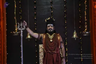 A bearded man with long dark hair and a gold crescent ornament stands before a dark textured backdrop strung with fairy lights and hanging brass bells. He wears a rust-red draped robe, a gold ceremonial necklace and dense rudraksha mala strands, and raises his right arm to hold a silver trident. A gilded chair stands beside him. The appearance-based label is a person; the image is a photograph of a darshan setting with the trident and bells as key props.