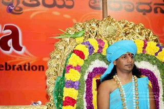 A seated man in a turquoise-blue turban and blue wrap, bare-shouldered, wears a red bindi and a long ceremonial necklace of gold beads and crystal. He sits on a gold ornate throne decorated with yellow marigolds, purple flowers, red roses and green leaves. A green parakeet perches on the throne's top edge. Behind him an orange banner shows large red lettering and Kannada script. a person: the long hair, ornate gold beads and saffron-toned royal setting are noted cues, though the appearance is only partly clear from this view.