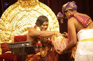 A photo of a ritual bathing scene in front of an ornate gold-leaf throne with a red velvet backdrop. A woman with wet hair, wearing a maroon and orange sari, a red and white flower garland and a gold necklace, sits on a red cushioned seat and smiles with eyes closed while water is poured over her head. A bare-chested priest in a purple turban and white dhoti with a gold border pours water from a decorated kalasha pot.