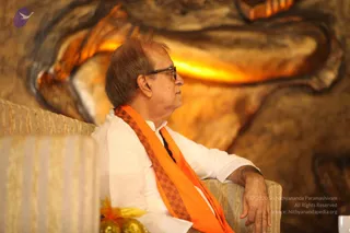 A seated man in a white kurta wears an orange-and-brown checked shawl draped over his shoulders and dark-rimmed glasses. He rests one forearm on the arm of a cream striped sofa and gazes to the side. Behind him a large glowing golden carved relief of an animal-like form fills the backdrop. Small gold-wrapped items sit at the lower left. The setting appears to be a formal stage or hall. Appearance is not sufficient to identify him.