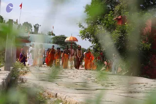 Outdoor photograph of a procession walking along a stone-paved path between a green tiled-roof temple structure and leafy trees. At the centre, a bearded man in a dark red-brown dhoti and a rudraksha-style beaded necklace with a decorative garland walks beneath a large ornate orange-and-gold parasol held by an attendant. Around him, several men in saffron-orange robes and dhotis walk in step, and a few in white dhotis and shirts follow. A woman in a purple sari stands at the left edge. Golden finials and red flags appear behind the buildings.