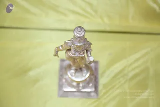 A small silver metal figurine of a dancing deity murti stands on a square silver pedestal against a yellow satin cloth. The figure has an ornate high headdress, decorated armlets and chest ornaments, and one arm extended outward while the other holds a small cylindrical object. No physical people are visible in the scene.