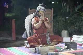 A seated man in a rust-orange sleeveless top and dhoti with a gold-edged sash and a leopard-print cloth wrap sits with palms pressed together in namaste at night. He wears a leopard-print headwrap, yellow tilak, rudraksha bracelets and a gold necklace, with long hair falling down his back. In front of him on a striped mat are stone mortars and pestles, stone bowls and a black stone block. A microphone stands at right, and a carved white throne-like chair with a dark background of trees sits behind him. The figure's identity cannot be confirmed from appearance, so no a person label is applied.