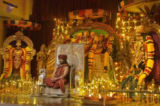 A seated man in a saffron-red robe and matching red turban sits cross-legged on a silver-framed platform in front of a golden shrine. He wears a red-and-beaded rudraksha mala and a gold-toned ceremonial necklace. A microphone stand stands beside him. Flanking him are deity murti with ornate gold and silver figures, decorated with yellow and orange marigold garlands. Rows of brass oil lamps and tall lamps glow on the floor in front of the platform, and fairy lights are strung along the backdrop, with colourful striped canopies overhead.
