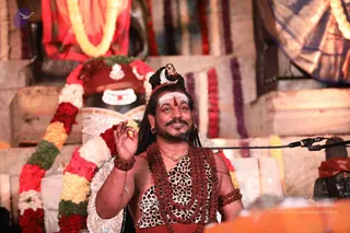 A man seated before a microphone on a stand, gesturing with his right hand raised in a mudra-like pose while speaking. He wears a white tilak with a red bindi on his forehead, a crown with a crescent ornament, rudraksha mala necklaces of brown beads over a leopard-print sleeveless top, a gold chain, bangles and a red waist cloth. Behind him stands a decorated Shiva lingam with a silver face and red-and-white garlands, flanked by floral malas in red, white, yellow and green, and draped fabric in red and blue. The setting suggests a discourse or satsang at a temple-like stage.