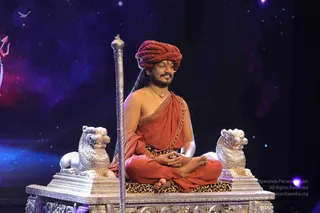 A man seated cross-legged on an ornate silver throne with two silver lion figures at its sides and a leopard-print cushion beneath him. He wears a red-orange turban, a red-orange shawl draped over bare shoulders, a gold chain necklace with a gold-and-beaded mala, and holds his hands in a meditation mudra. A tall silver staff stands beside the throne. The backdrop is a dark starry space with a faint red trident motif at the left edge. Swamiji (probable): the seated man shows long swept-back hair, a red-orange turban, orange shawl and ceremonial gold necklace on a throne, a compatible presentation.