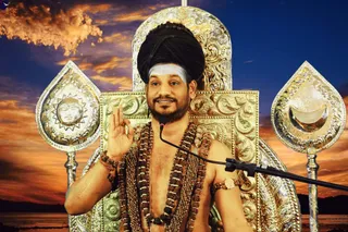A seated man in a silver-and-gold ornate throne with sun and crescent moon finials, a large black turban, a white vertical tilak on his forehead, and a beard. He is bare-chested, wearing layered rudraksha malas and gold chains, a beaded bracelet and an armband. He raises one hand in a blessing gesture before a microphone on a stand, with a sunset sky and sea behind him. a person: the black turban, bare-chested ceremonial dress, rudraksha malas and gold ornaments match the page's a person reference, but the photo is a composite with a painted background, so the identity is only appearance-based. No other people are visible.