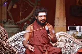 A bearded man with long dark hair and white tripundra markings on his forehead and arms sits on an ornate silver throne-style chair, speaking into a handheld microphone. He wears a rust-red ochre robe with a gold-bordered shawl draped over one shoulder and a rudraksha-style mala across his chest. A small red thread is tied on his wrist. Behind him stands a bronze Nataraja-style dancing deity sculpture and a red-painted wall with a stone pillar. A leopard-print cloth is visible at the left edge.