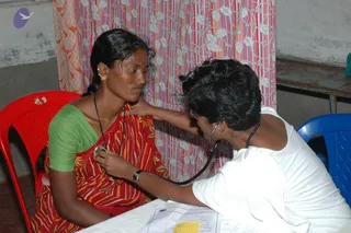 A medical check-up at a free medical camp. A woman in a red and yellow striped sari with a green blouse sits on a red plastic chair, with a red bindi and gold earrings, while a health worker in a white top and rudraksha-style beaded necklace listens to her chest with a stethoscope. A pink and white leaf-patterned curtain hangs behind them, and a form lies on a white table.