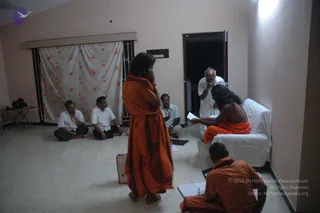 Indoor photo in a plain room with white walls and a speckled floor. A white curtain with red flower print hangs at a window on the left. Five men in white shirts or white dhotis sit on the floor or stand near the back, several with hands folded in greeting. A central figure in orange robes stands in front, one hand raised to the face. A long-haired figure with bare upper body and orange cloth sits on a white-covered chair at right, holding a sheet of paper. In the lower right, a person in orange robes sits on the floor with a notebook and a tablet. A camera bag and a small table sit at the far left. The scene looks like a private meeting or consultation with devotees.