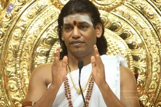 A man seated in front of an ornate gold repoussé backdrop speaks into a small microphone, both hands raised with palms facing inward in an expressive gesture. He wears a plain white dhoti-style cloth draped over his shoulders, a gold chain necklace, and multiple strands of rudraksha beads. He has long dark hair swept back, white vibhuti (sacred ash) horizontal lines on his forehead, and a red kumkum dot between his brows. A rudraksha bracelet is visible on his wrist. The setting suggests a discourse or satsang. Swamiji (probable) is shown in person, with long swept-back hair, white forehead markings, rudraksha malas and a gold necklace.