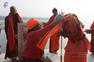 Outdoor riverside scene on a stone platform by a wide, pale river under hazy winter light. A man in an orange turban-style cap and orange-red robes kneels and drapes an orange cloth over the head of an older bearded sadhu in orange and beige robes who bows forward, a silver trident-topped staff (trishul) planted beside him. Behind them, robed men in orange and saffron stand watching, one in a dark knit cap and another holding a hand near the scene, with a silver ornate panel and a water bottle on the platform. Ritual blessing or honouring of a sannyasi at the riverbank.