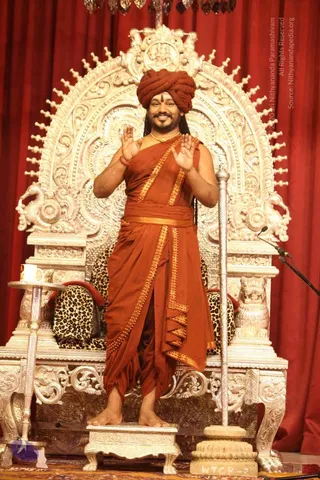 A man stands on a silver-toned footstool in front of an ornate silver throne with a leopard-print cushion, against red drapery. He wears a rust-orange turban, orange-saffron draped robes edged with gold-toned trim, a gold chain, and a red-tinged wrist thread, with a vertical tilak on his forehead. He raises both palms toward the audience in a blessing gesture. A silver staff stands beside him, a microphone is at right, and a silver tray with a cup sits at left. a person by appearance cues: long hair, turban, saffron ornate clothing, gold necklace, seated-throne setting, with the page noting his presence.