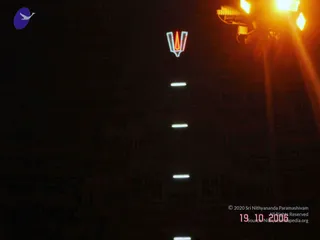 A dark night photograph of a building facade with a glowing neon-style trident (trishul) sign at the top, lit in red and white with a flame-like orange centre. A row of white fluorescent or tube lights runs down the wall beneath it, and a bright orange floodlight glares in the upper right corner. No people are visible; the scene is architectural and illuminated, with a faint carved or pillared wall barely visible below.