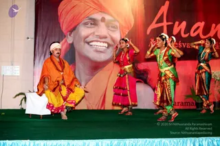 A stage performance of classical Indian dance under a large backdrop printed with a smiling man wearing an orange turban and orange robes, with the word "Anan..." in red script. A seated male performer in an orange kurta over yellow dhoti with pink border and a white head cloth sits at left, hands raised, beside a white-draped stool. Three female dancers in colourful costumes stand in dance poses: one in pink and red with a golden waist belt, one in green and red with a gold belt and jewelled necklace, and one in teal and red. The stage has a green floor covering and potted plants at the edge.