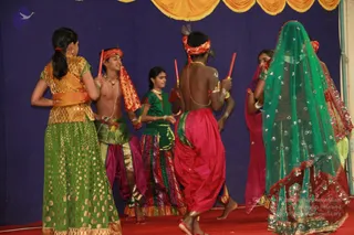 A stage performance of a folk-style dance by young performers in colourful traditional costumes against a deep blue backdrop with a gold-fringed valance. A young man in the centre, bare-chested with a red and gold headband and dhoti-style red trousers, holds a short red stick. Beside him, a boy in a red headband and pink trousers holds a red stick, and a girl in a green blouse and striped skirt stands among them. On the right, dancers in green and pink veils and embroidered outfits move across a red stage floor. A girl in a green and gold lehenga stands in the left foreground with her back to the camera.