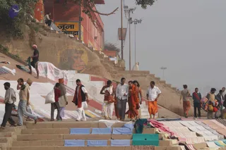 Several people walk down a stone ghat staircase beside the Ganges in Varanasi on a hazy morning. A red-clad figure in a saffron-orange dhoti and shawl leads a group, with a man in white kurta and grey trousers walking beside him. A bare-chested man in white dhoti carries a brown bag, and a man in an orange dhoti walks behind. Cloth is spread to dry on the steps, and a faded painted monkey face decorates the wall above. Signage reads Juna Akhara on a red building. Visible physical people number about eight to ten.