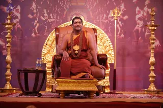 A man with short dark hair sits on an ornate brown leather throne with gold-carved arms and a golden arched backrest in front of a pink-lit backdrop of dancing figures. He wears a saffron-orange robe with a wrapped shawl, a rudraksha mala with a large ceremonial gold pendant, and a red bindi. His hand rests on the armrest. Beside him stand tall gold lamp stands and a gold trident, with a small wooden side table holding water bottles. The a person candidate is shown in person; appearance cues are saffron robes, rudraksha mala and ceremonial gold pendant, with throne and dais setting. Two people are visible in the foreground edge.