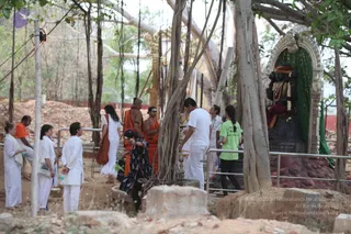 A photograph of an outdoor ashram tour under a large banyan tree with hanging aerial roots. Several visitors in white clothing stand on sandy ground behind a metal railing, including a woman with a white shawl and a bag. Two monastics in orange and saffron robes stand near the centre, one bald with bare shoulders and a shawl, the other with a dark shawl. A man in a white shirt and a woman in a green t-shirt look toward a black stone deity murti in a carved arched niche, adorned with a red and gold cloth, a garland and a beaded mala. Red earth and construction mounds surround the area.
