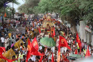 A large street procession moves through a crowded roadside lined with tents, stalls and shade trees. At the centre, a gold-topped ornate palanquin with floral garlands carries a seated figure in saffron-coloured clothing, with a bare-chested attendant standing nearby. A green umbrella with mirrored embellishment shields the carriage, and uniformed police in khaki and officers in yellow vests guide the route behind yellow barricades. Many devotees in white dhotis and orange shawls, some wearing rudraksha malas, walk alongside, while red flags bearing a portrait of a saffron-clad sannyasi wave above the crowd.
