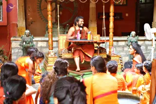 A seated man in a rust-red wrap and a saffron-brown shawl sits on an ornate gold-and-wood throne with a small table holding a microphone, water bottle and cups, speaking and smiling in a courtyard. Behind him are a red wall, a dark bronze relief and two green-patinated standing figures. Around him, women and girls in bright orange and yellow saris sit on the floor facing him, several with hair in buns and flowers; a woman in orange sits to his right. A white pedestal fan stands at the right edge. Swamiji (probable): the seated man has long swept-back hair, a beard, a shawl and a throne, consistent with the page's reference to a discourse on Ganesh Chaturthi.